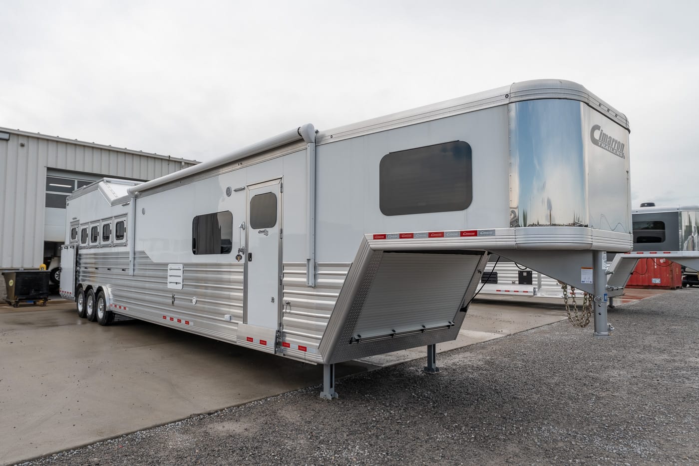 Silver aluminum horse trailer with living quarters parked beside a building; gooseneck hitch and folded loading ramp visible to the right.