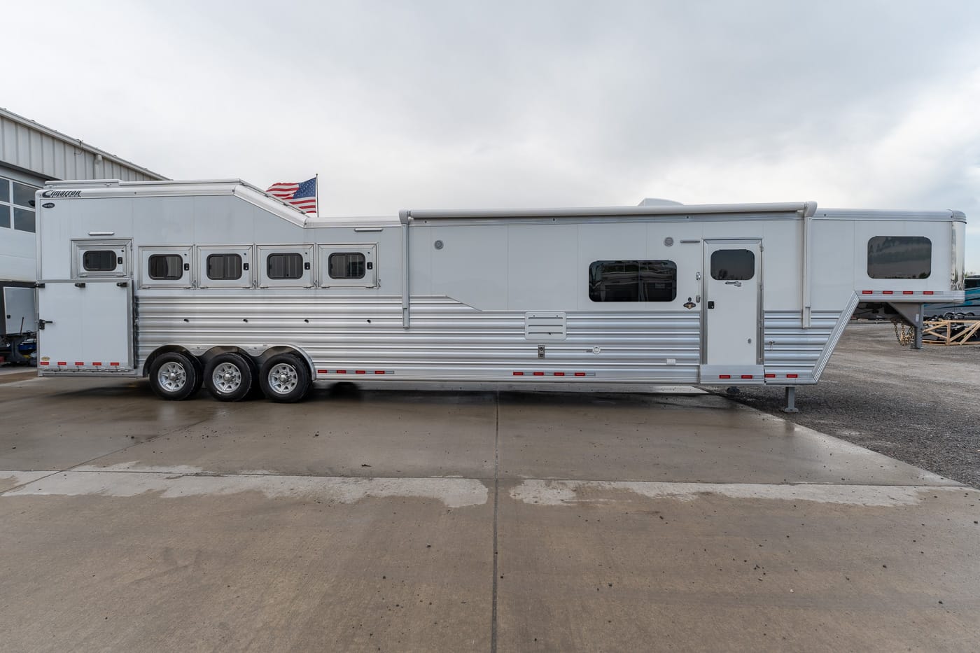 Triple-axle silver horse trailer with living quarters, parked on a wet concrete lot with an American flag in the background