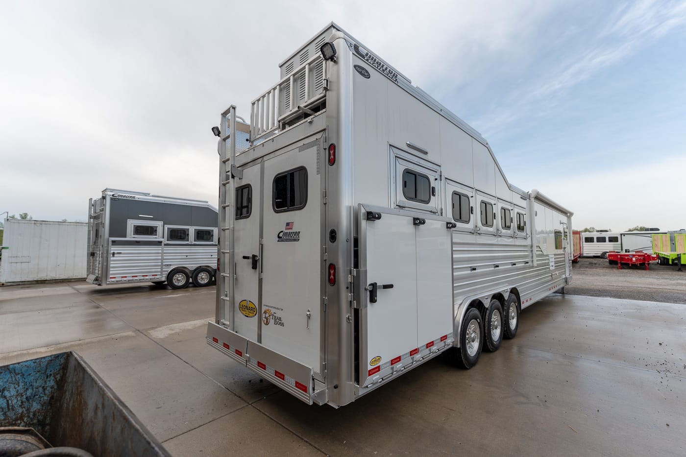 Silver multi-axle horse trailer with several windows and storage bays, parked in a trailer yard.