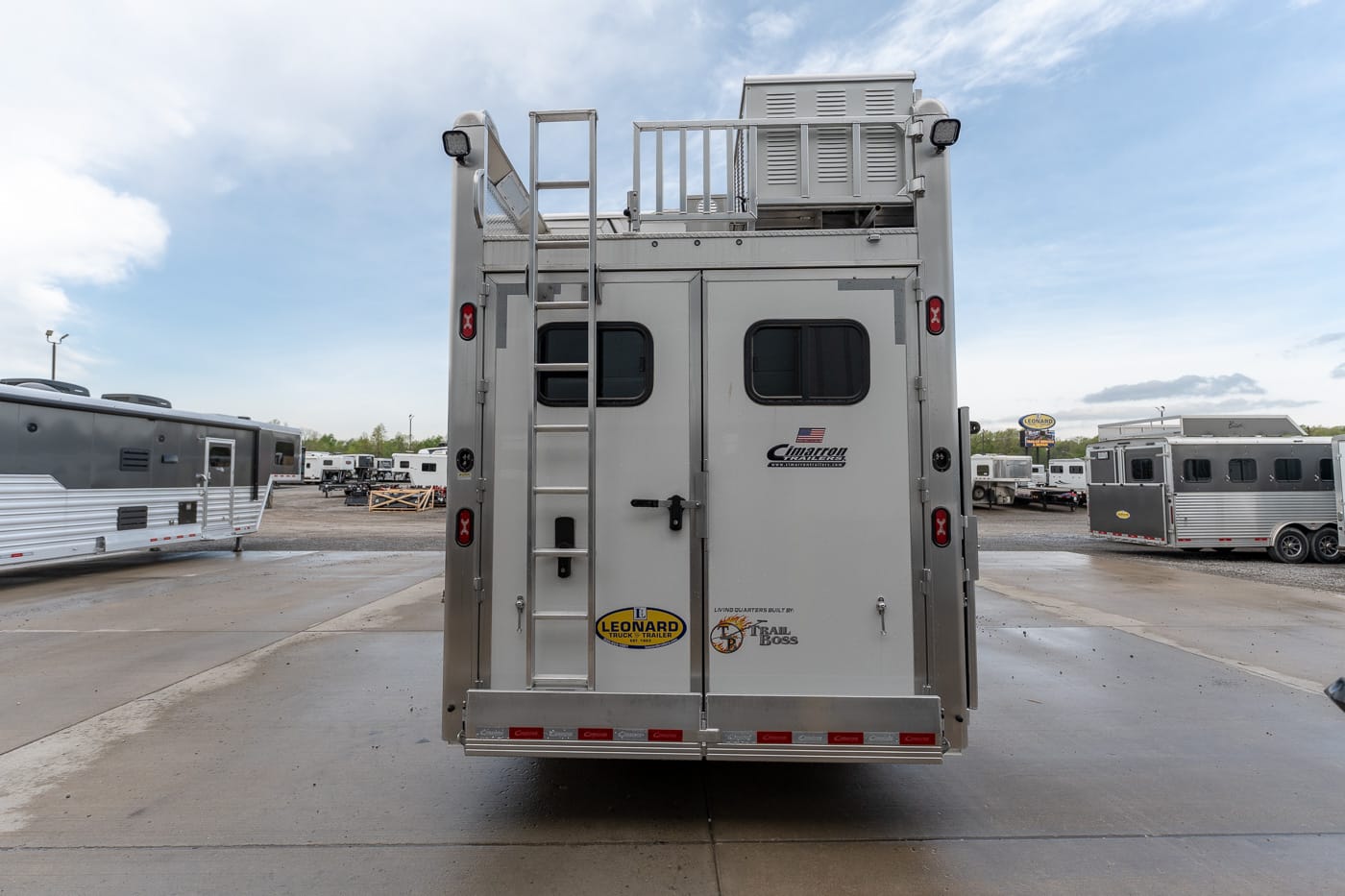 Rear view of a large silver horse trailer with a ladder, two small windows, and decals, parked in a trailer lot under a blue sky.