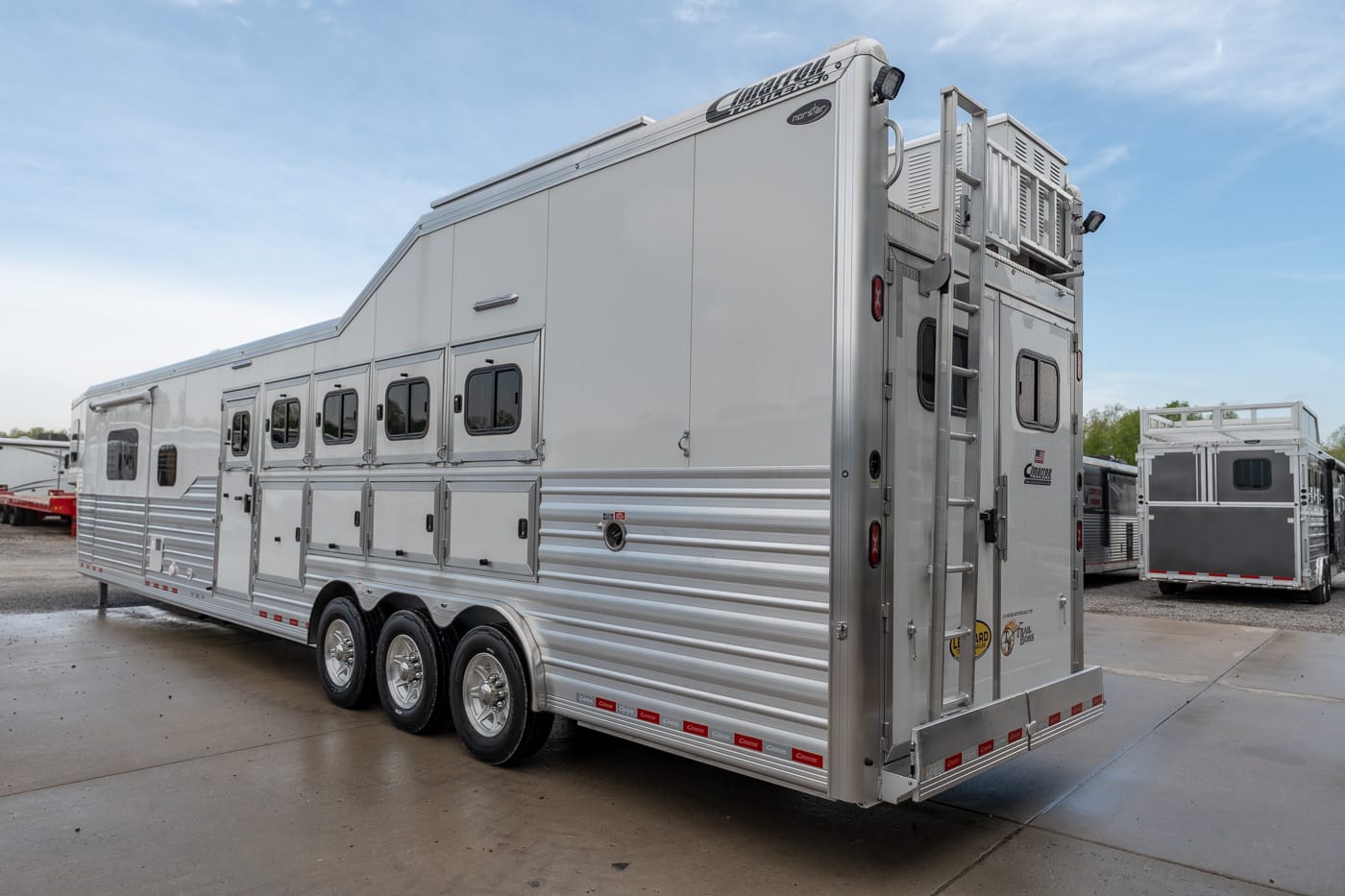 Silver aluminum horse trailer with three axles and multiple side windows, parked on a wet concrete lot (used for transporting horses).