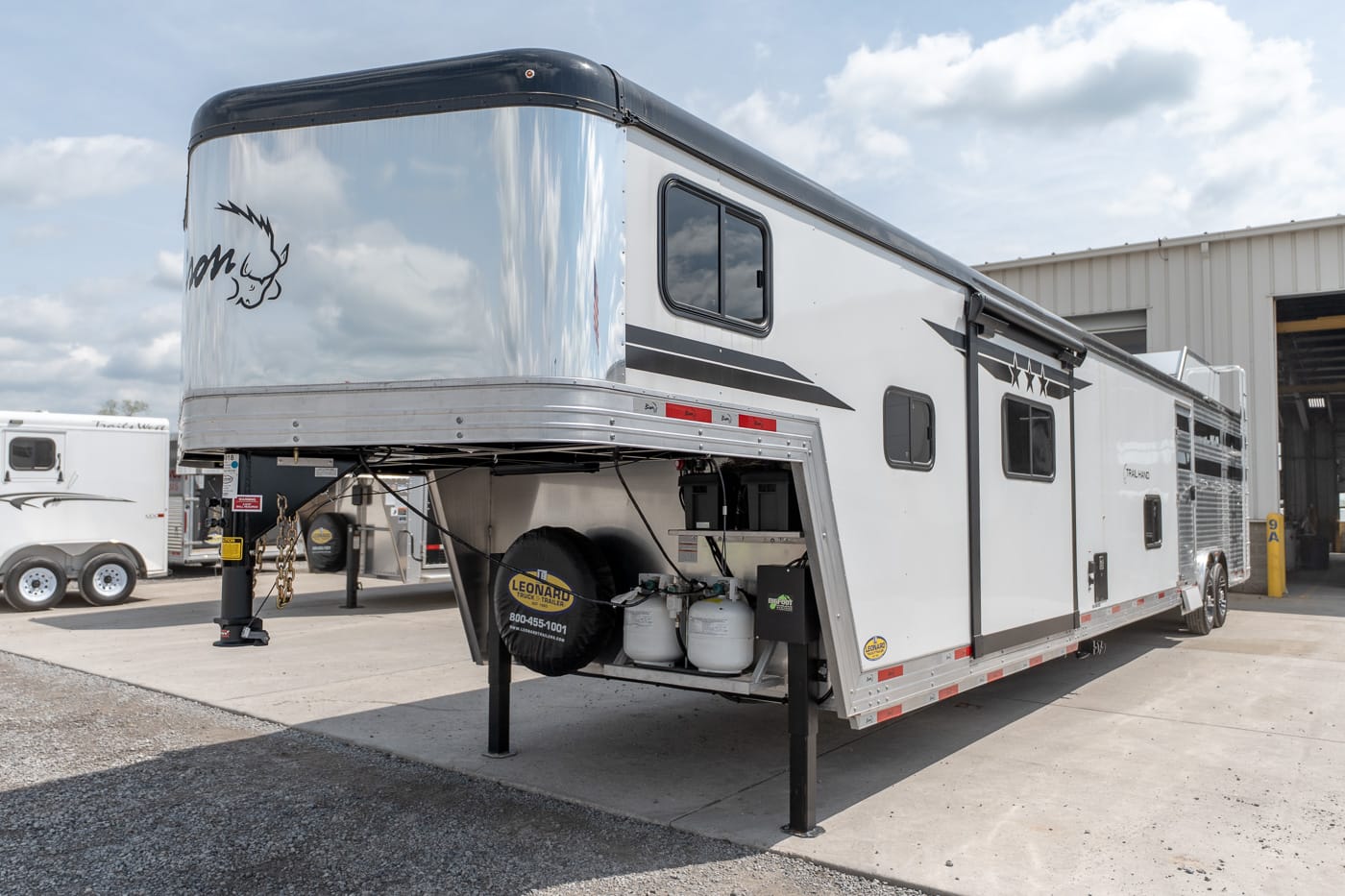 White fifth-wheel horse trailer with a raised front, parked on concrete with propane tanks and a spare tire beneath the hitch.