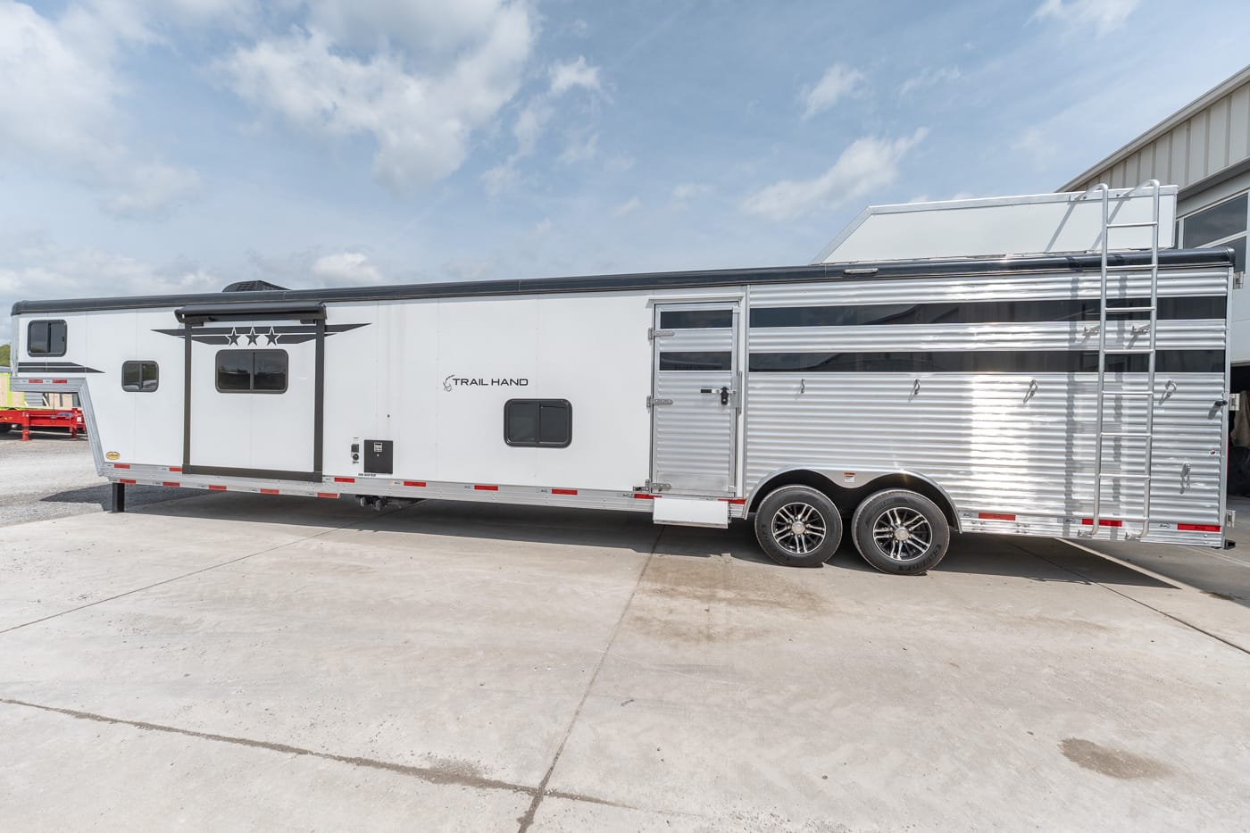 White horse trailer with chrome wheels and aluminum siding parked on a sunny concrete lot, side exit door visible and Trail Hand branding on the side.