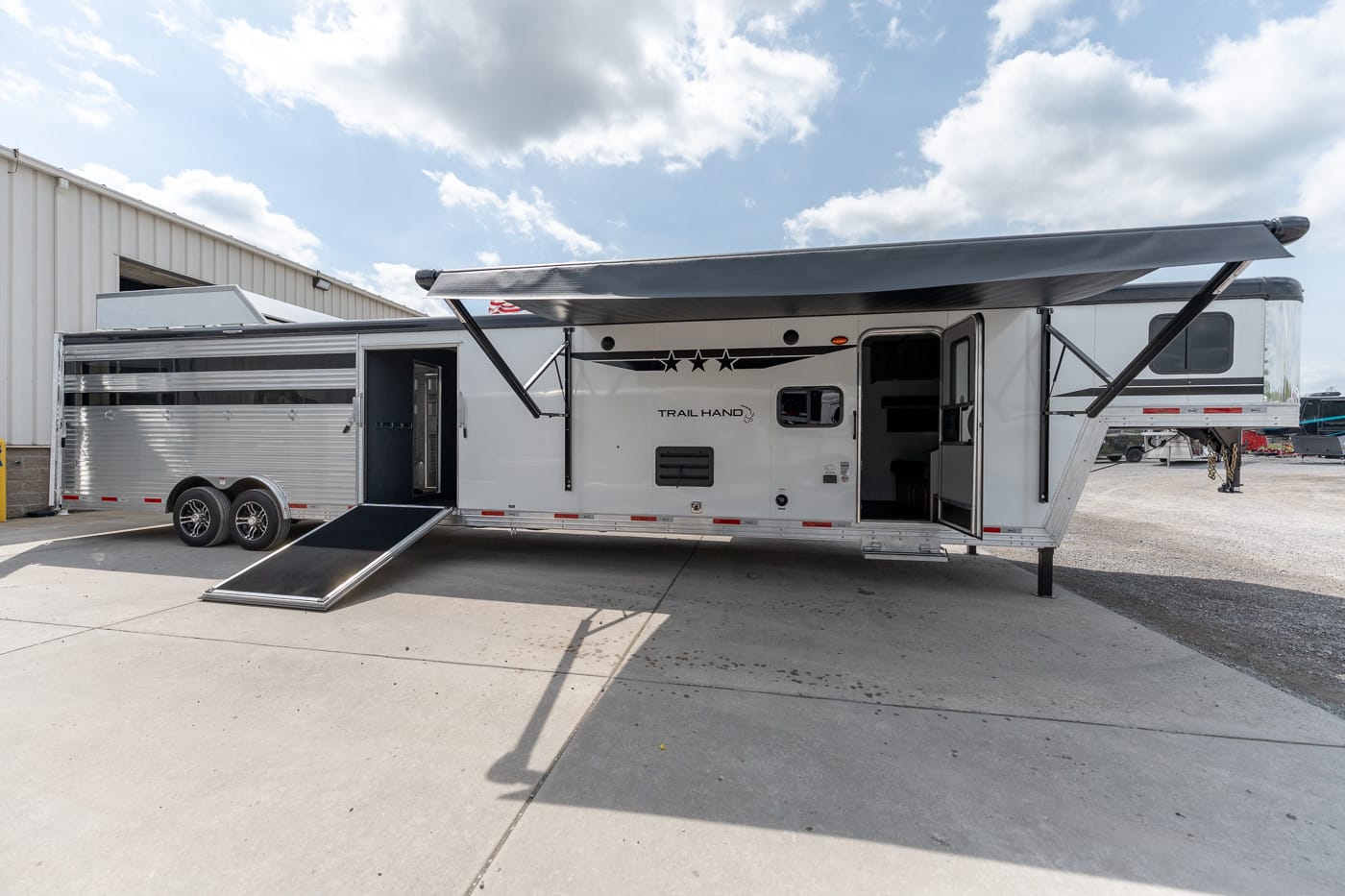 White and silver travel trailer with an extended awning and lowered ramp, parked on a concrete lot.