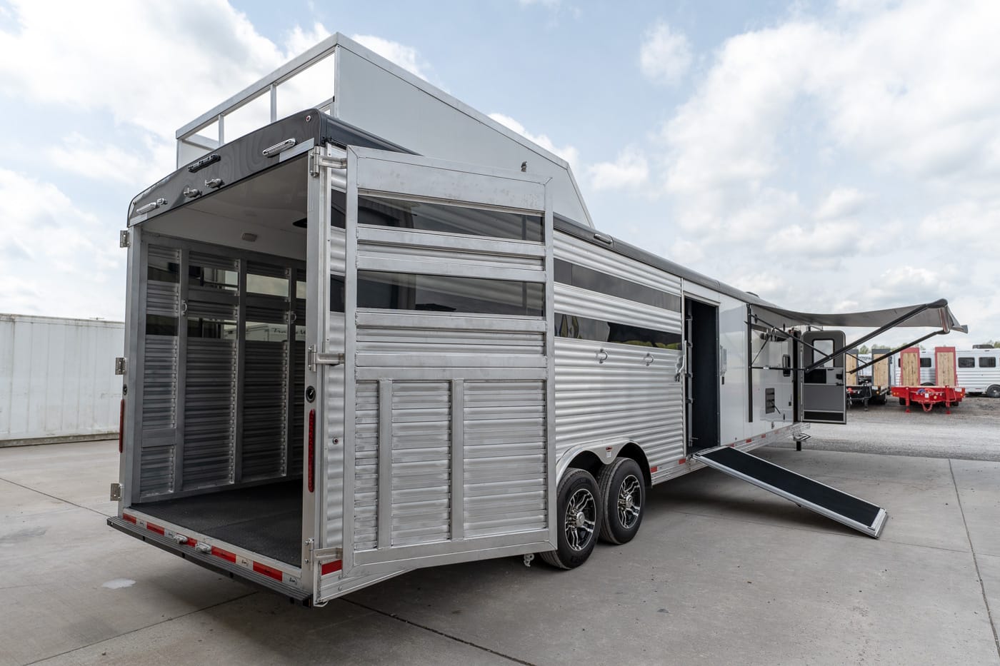 Silver horse trailer with rear and side doors open, ramp lowered, parked on a concrete lot under a blue sky with clouds