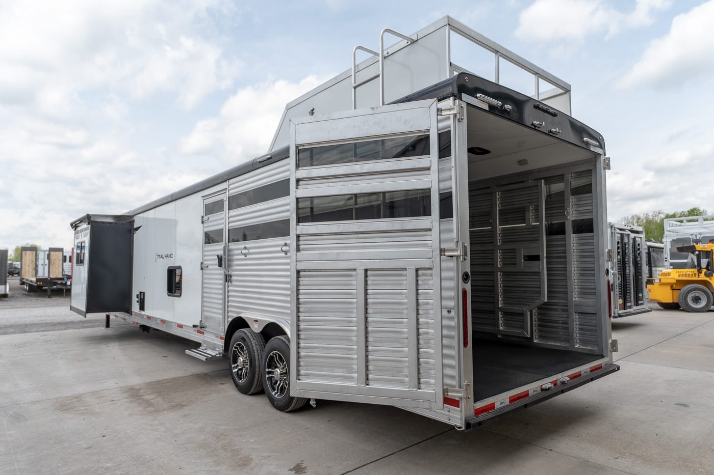 Silver aluminum horse trailer with multiple stalls and a down/open rear ramp on a concrete lot, cloudy sky above.