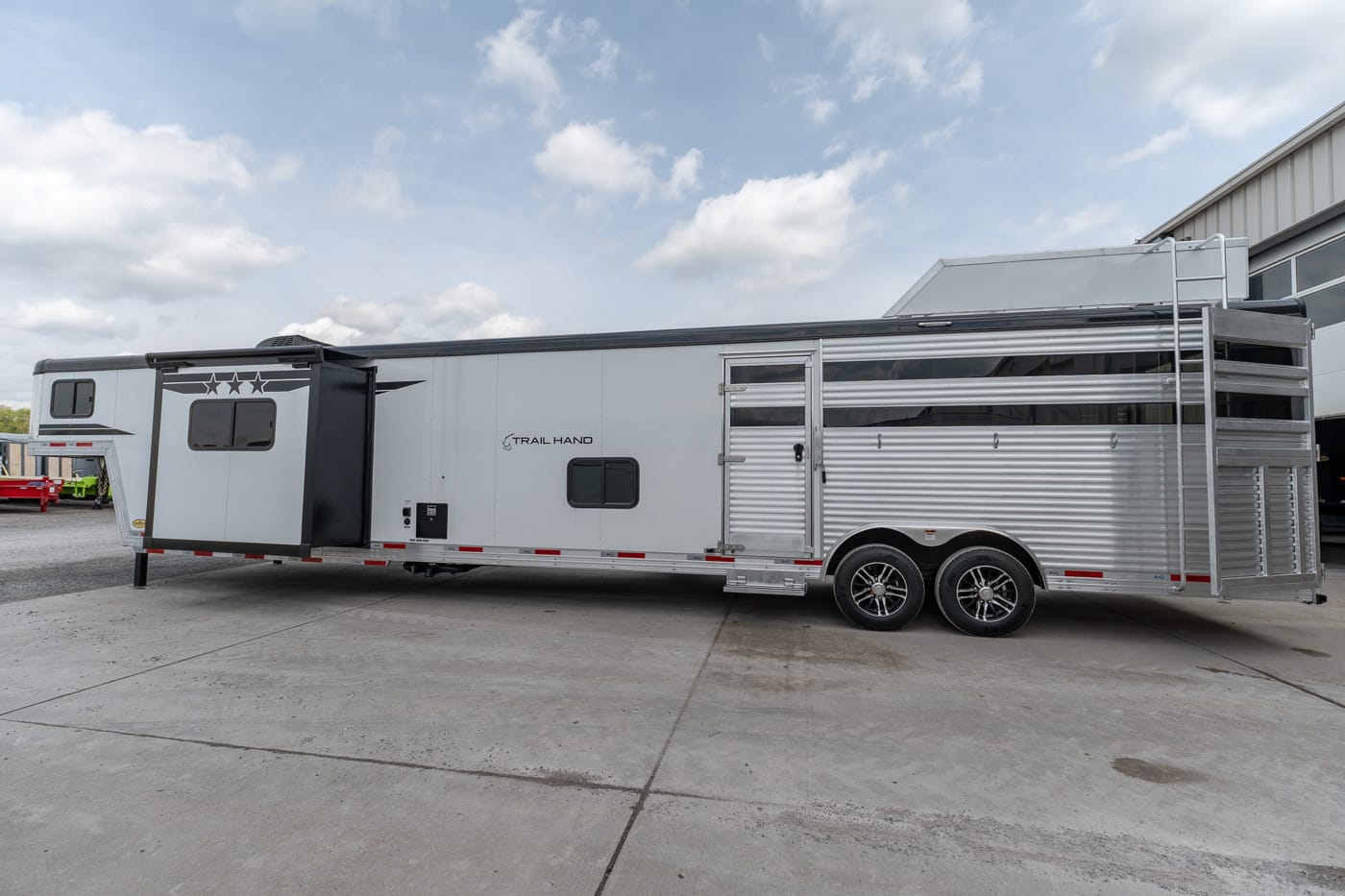 Long silver horse trailer with dual axles parked on concrete, hitch extended, outside a warehouse.