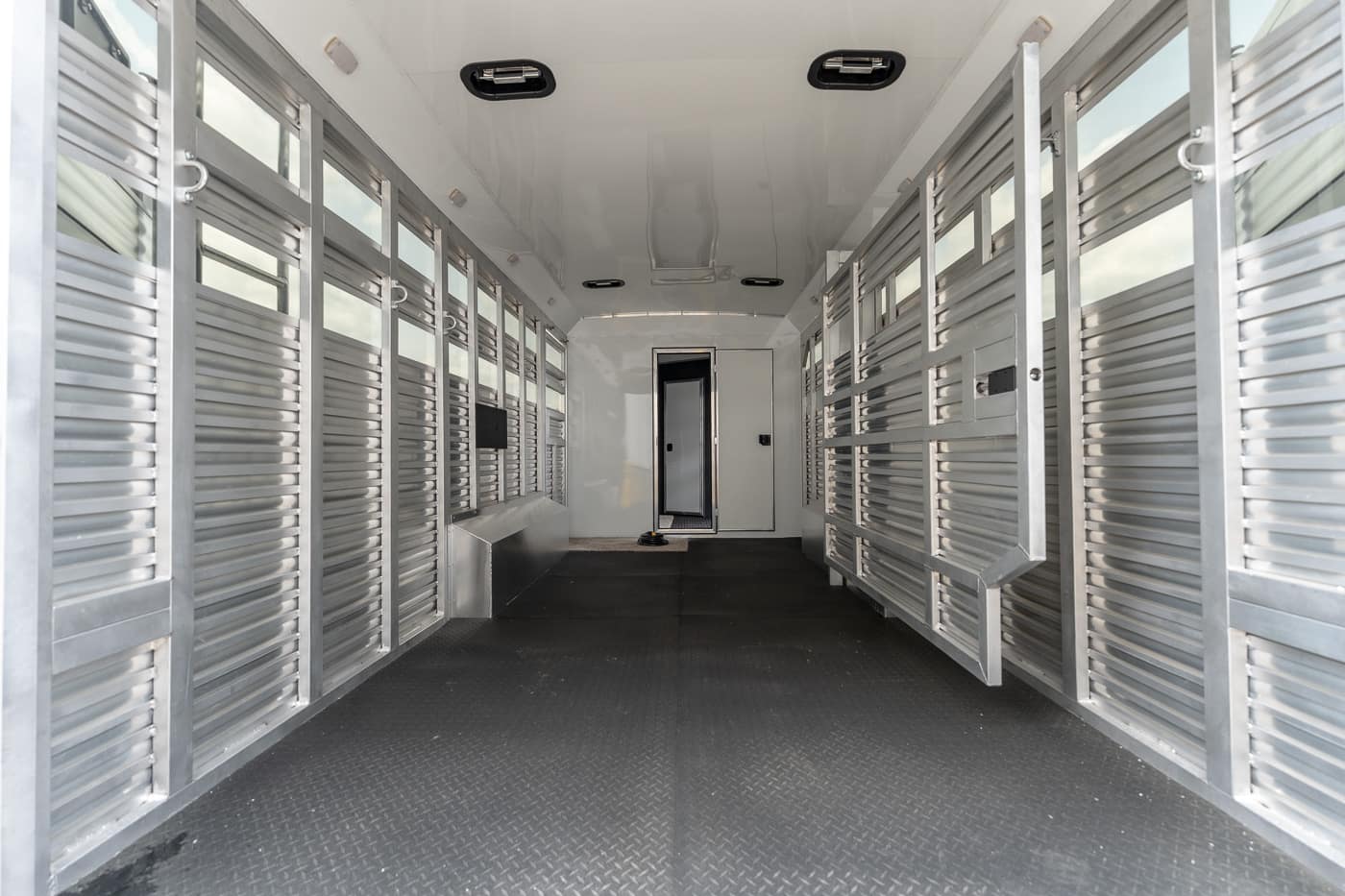 Interior of an aluminum livestock trailer with metal stalls along both walls and a central aisle, door at the far end.