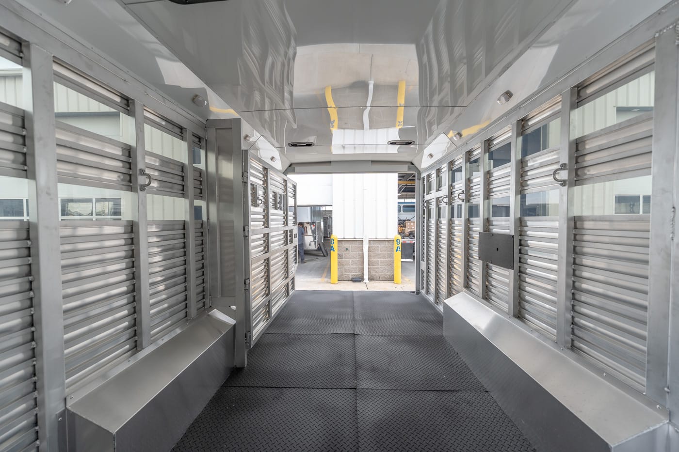 Interior of a silver livestock trailer with aluminum stalls on both sides and a diamond-plate floor, leading to an outside loading area.