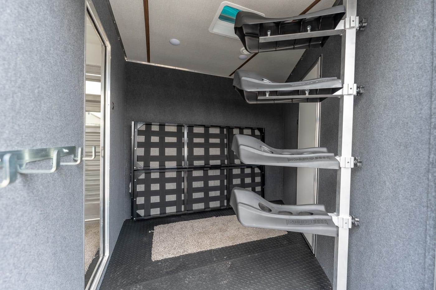 Inside a horse trailer with multiple gray saddle racks mounted to the right wall and a padded, rubberized floor area.