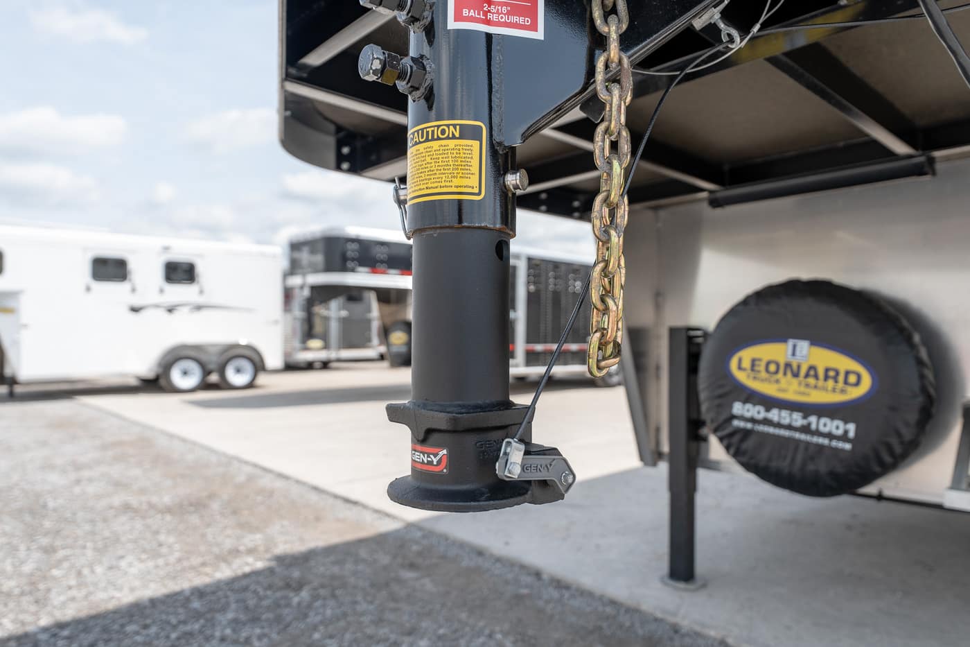 Close-up of a trailer landing gear jack with safety chain, warning stickers, and a Gen-Y label; spare tire with Leonard logo in the background.