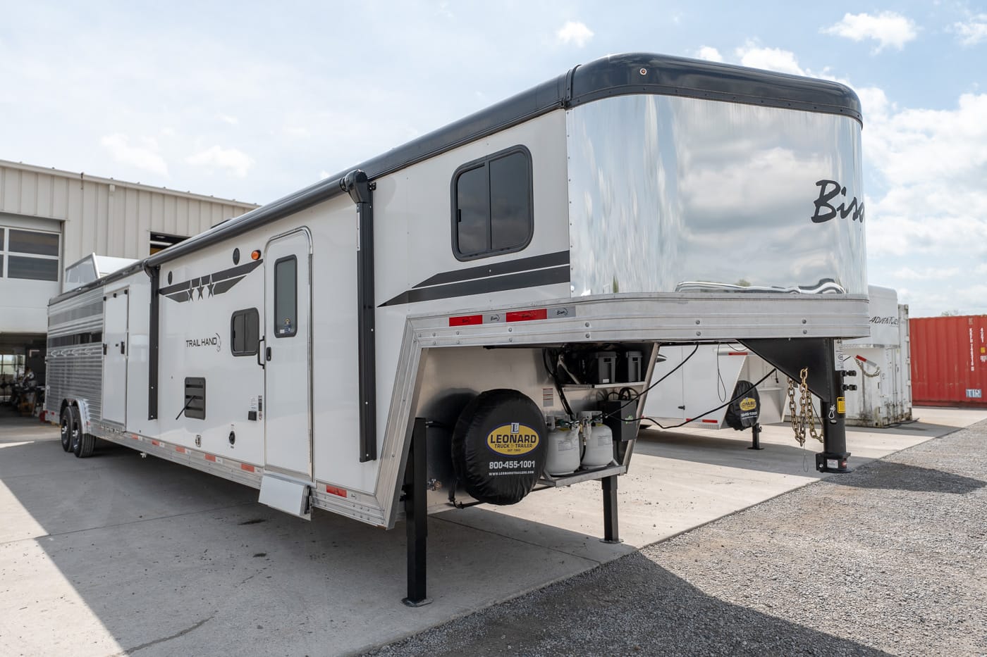 White gooseneck fifth-wheel RV trailer parked on a concrete lot, stabilizing jacks down and a spare tire mounted on the front.