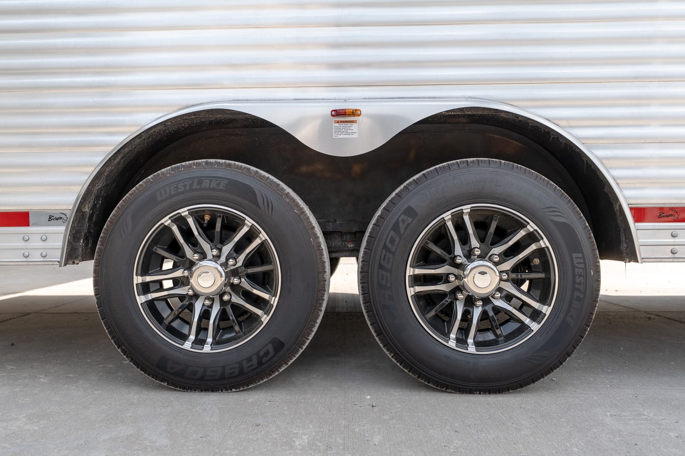 Twin-axle trailer with chrome-rimmed black alloy wheels and Westlake tires beneath a silver corrugated body, with curved wheel wells overhead.