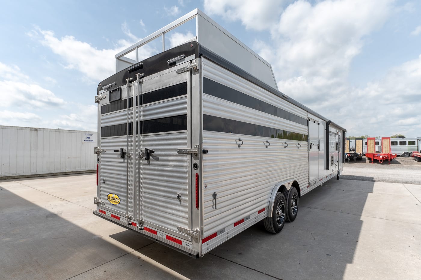 Two-axle silver aluminum horse trailer with rear double doors, parked on a concrete lot under a blue sky.