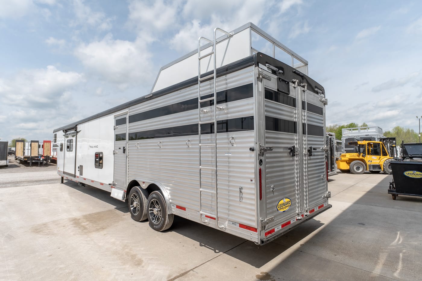 Aluminum horse trailer with dual axles parked on a concrete lot, rear double doors and a ladder on the back wall, branding near the bumper.
