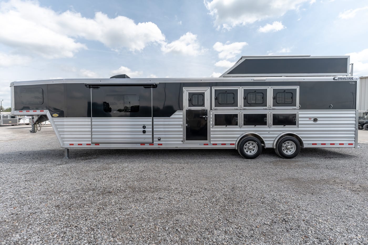 Side view of a two-tone aluminum horse trailer with multiple stall windows, parked on a gravel lot on a sunny day