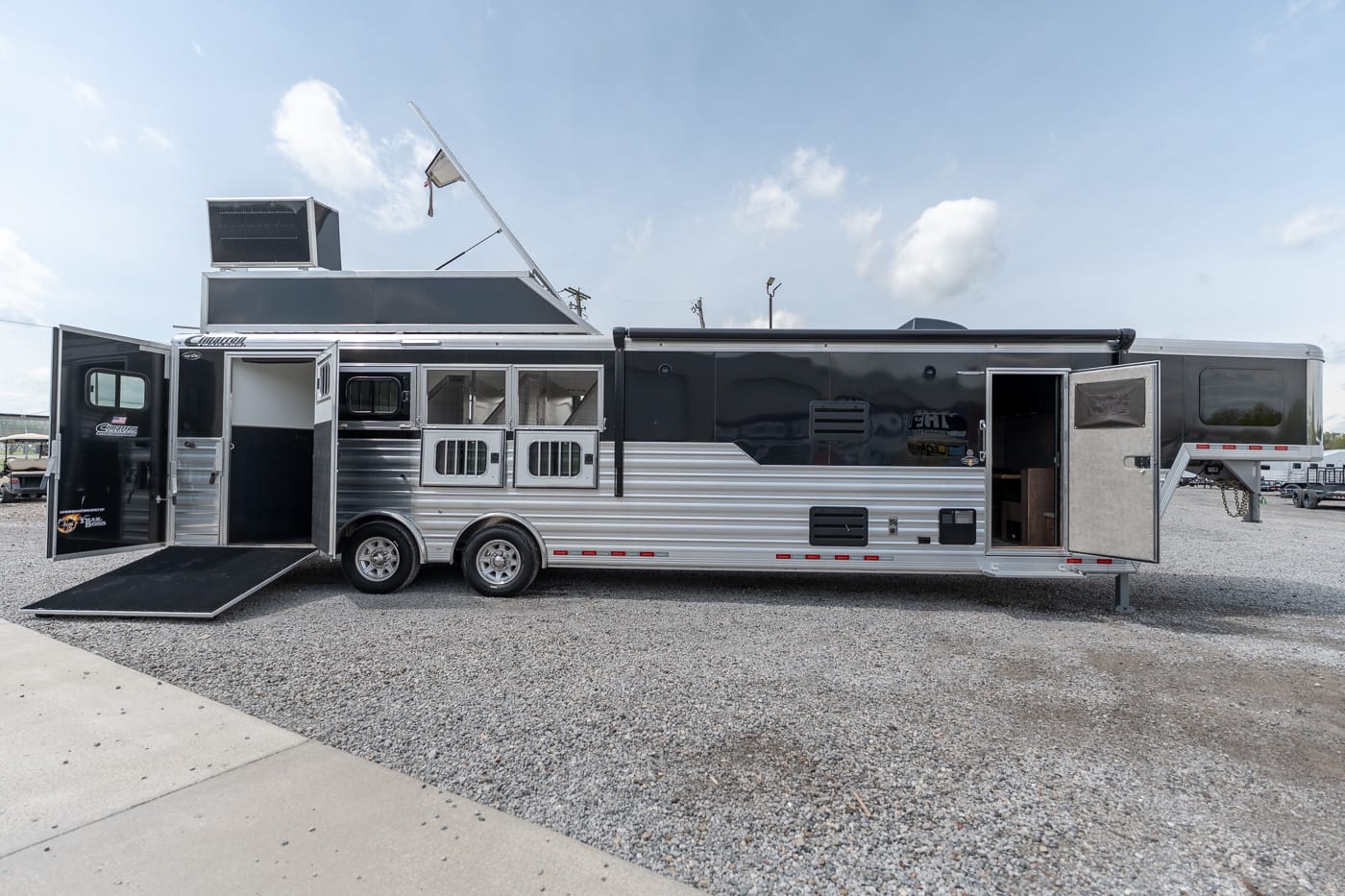 Long white-and-silver horse trailer with two axles and a side ramp lowered, living-quarters door open on a gravel lot; rear entry open.