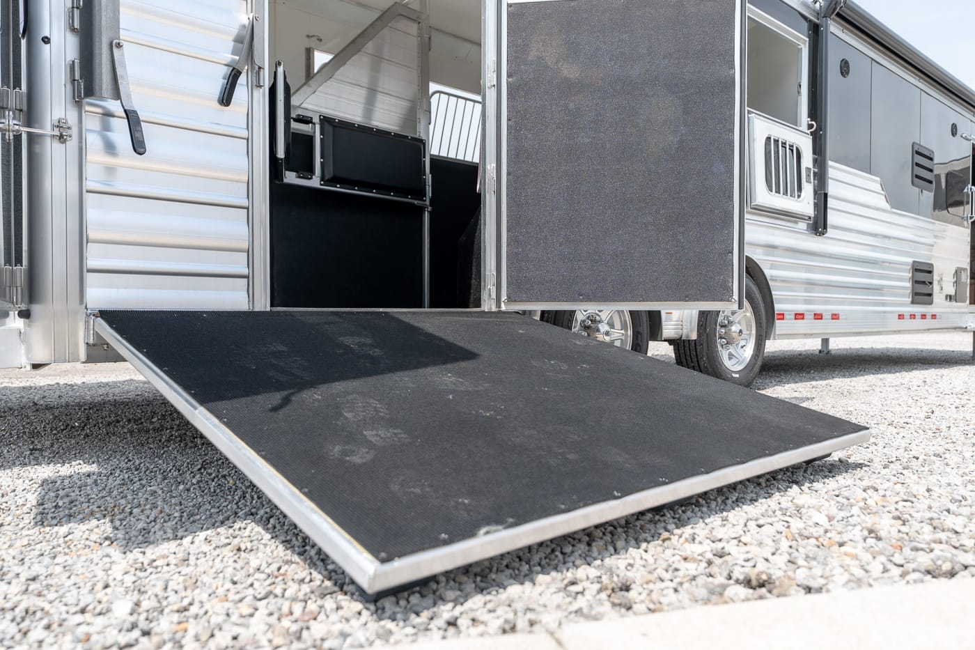 Open silver horse trailer with its loading ramp lowered onto a gravel surface, exposing the interior stalls and stairs inside the trailer.
