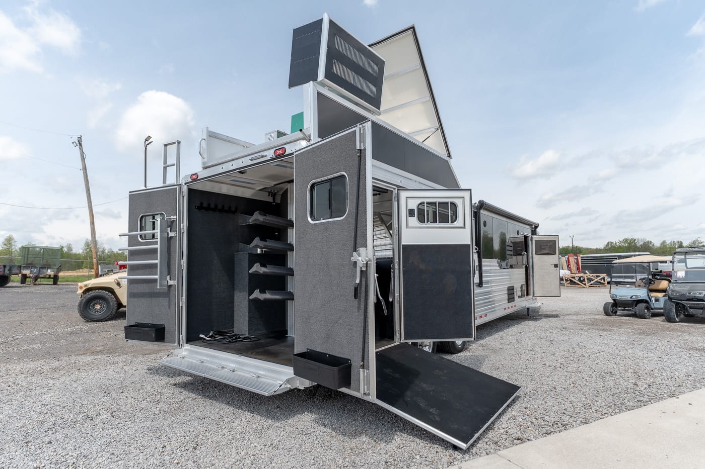 Open utility trailer with multiple storage compartments and a lowered rear ramp on a gravel lot.