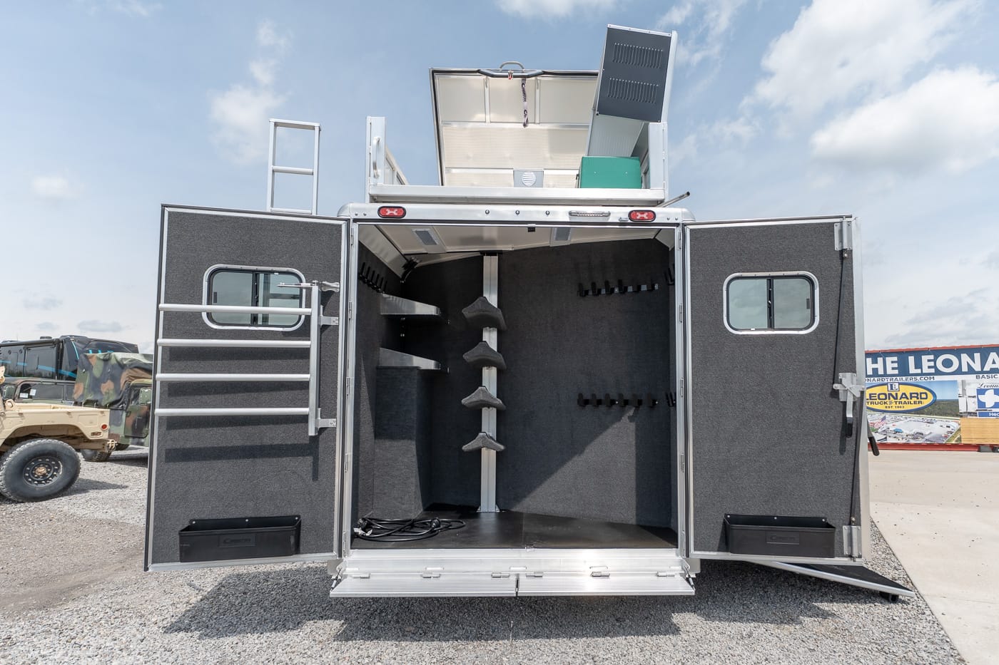 Rear view of a large silver trailer with its doors open, showing interior saddle racks and numerous hooks for tack on a gravel lot. The blue sky is overhead.
