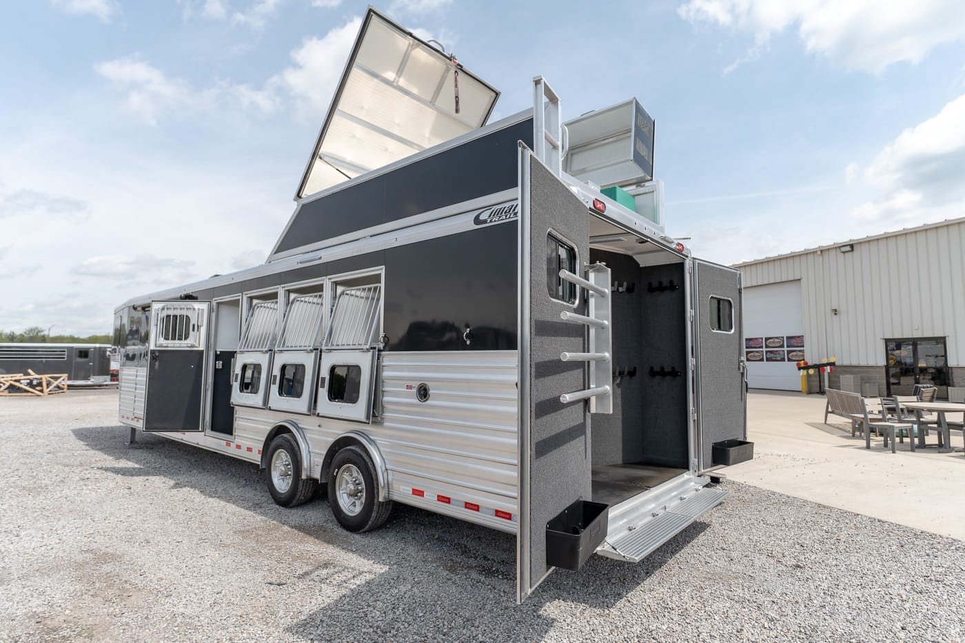 Large aluminum horse trailer with open rear ramp and tack compartments on a gravel lot under a blue sky