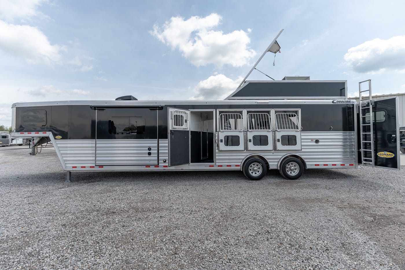Long aluminum horse trailer with side living quarters and open doors, parked on a gravel lot under a blue sky.