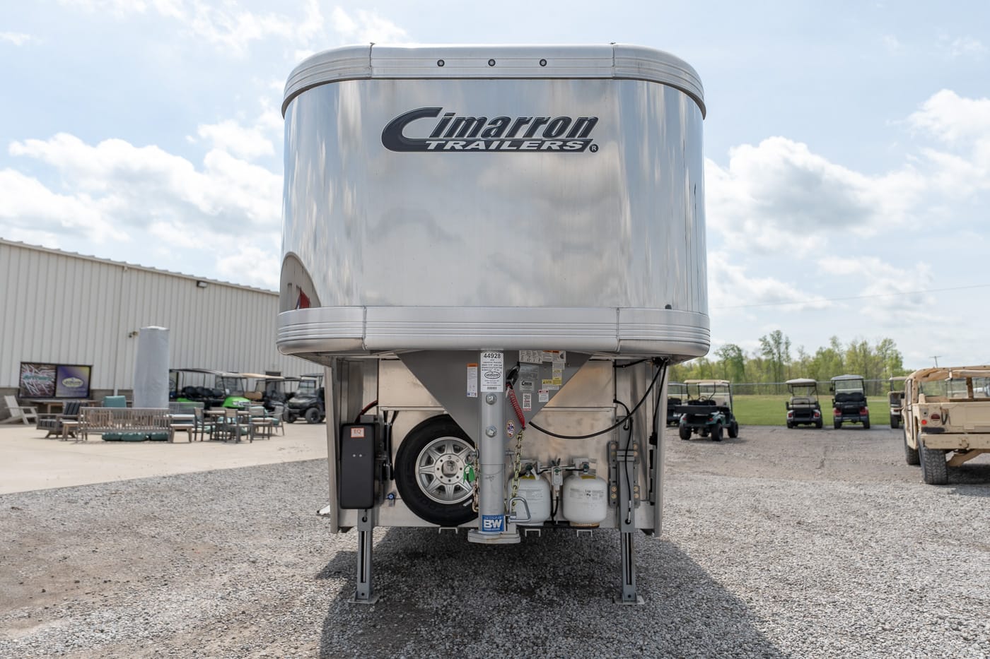 Back of a shiny aluminum Cimarron Trailers gooseneck trailer on a gravel lot, with spare tire and propane tanks visible at the hitch area.