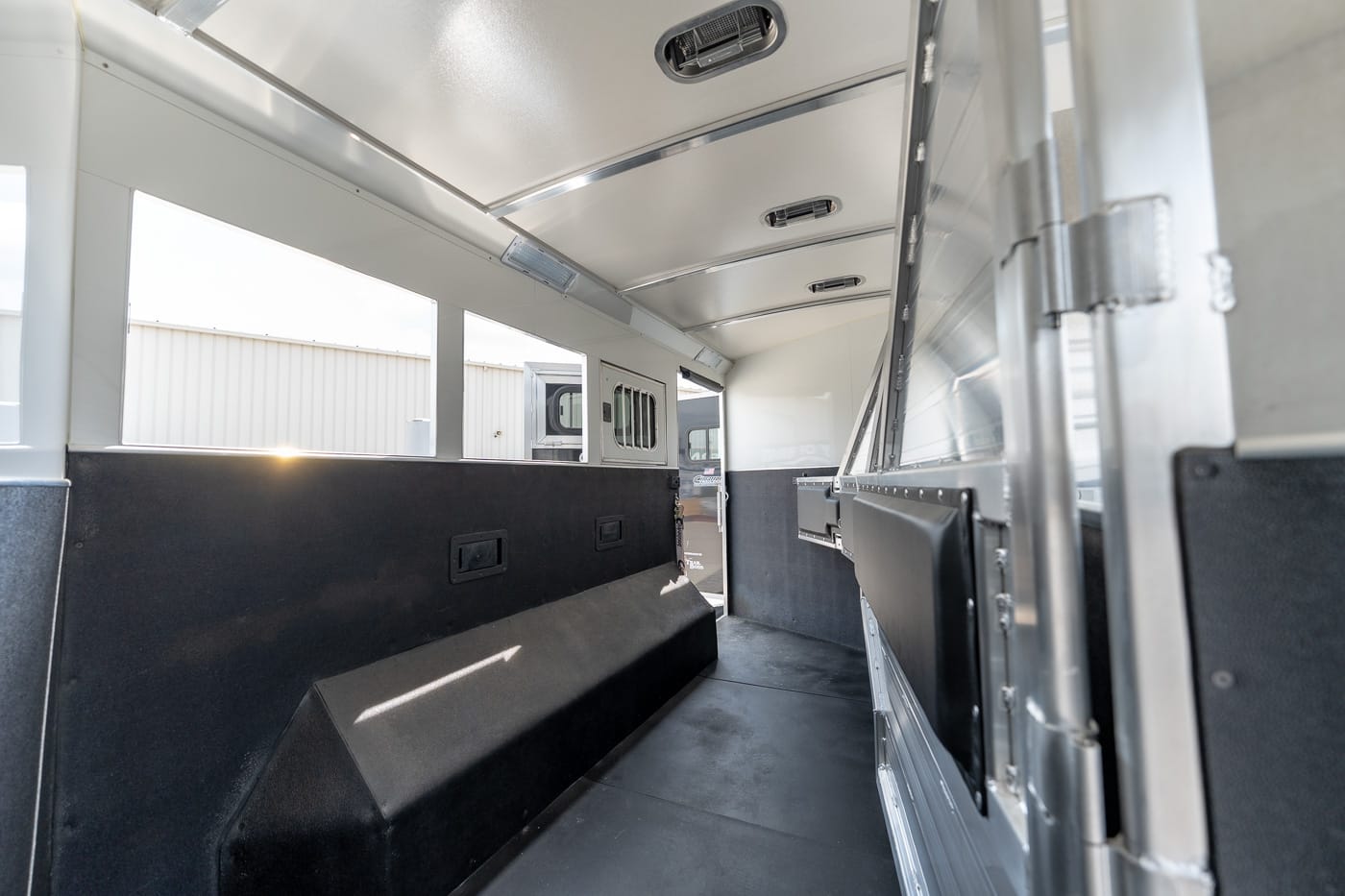 Interior of a light-filled aluminum trailer with black padded side benches and windows along the top as seen from the middle toward the rear door. 0