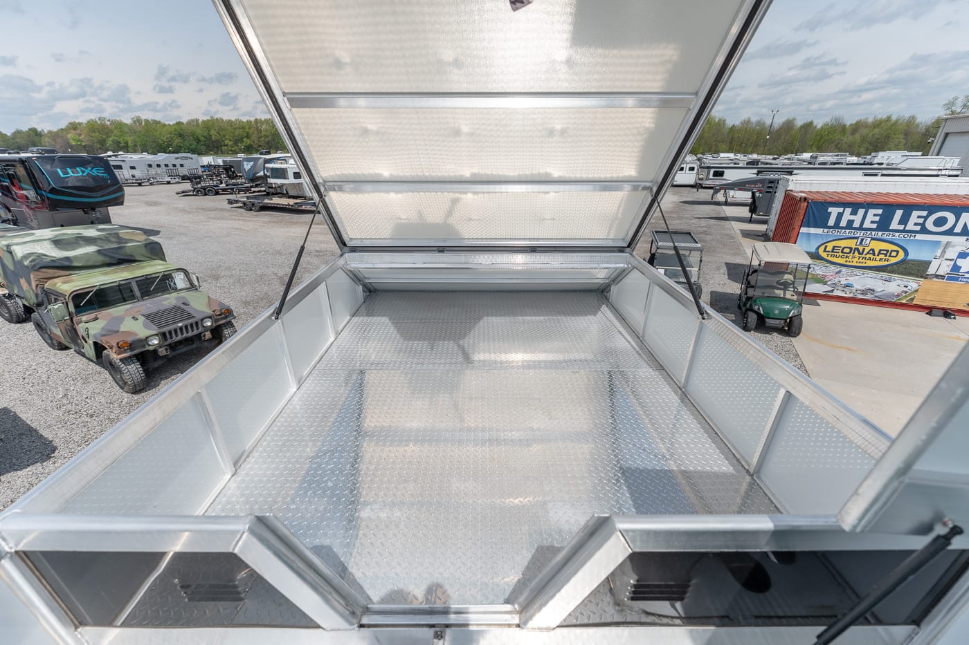 Open diamond-plate trailer bed with raised lid, empty and ready to load, on a dealership lot with trucks and trailers in the background.