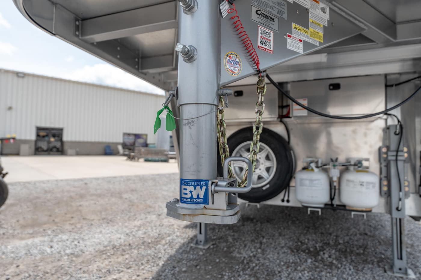 Close-up of a gooseneck trailer hitch with safety chains, a spare tire, and two propane tanks in the background.