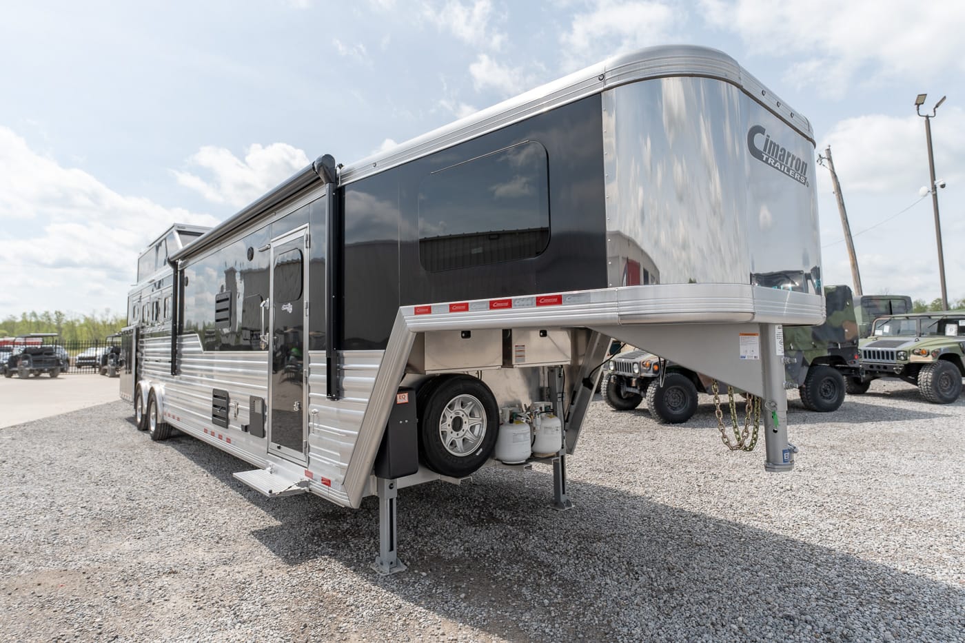 Side view of a large silver fifth-wheel travel trailer parked on a gravel lot, showing the raised front hitch, dual axles, and external compartments.