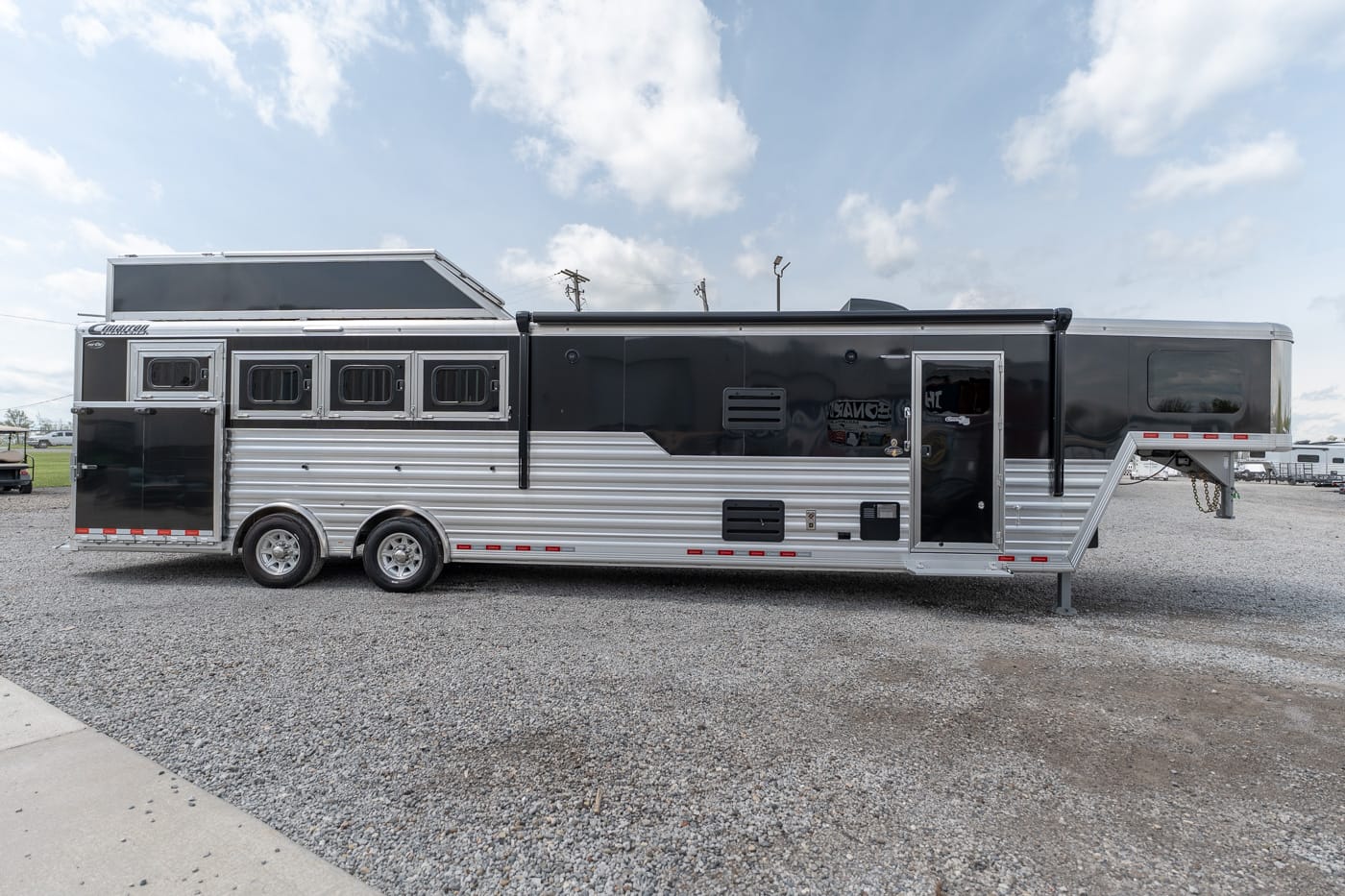 Long black-and-silver horse trailer with living quarters, mounted on a gravel lot, featuring multiple windows, dual rear axles and a raised front entry with stairs at the back end