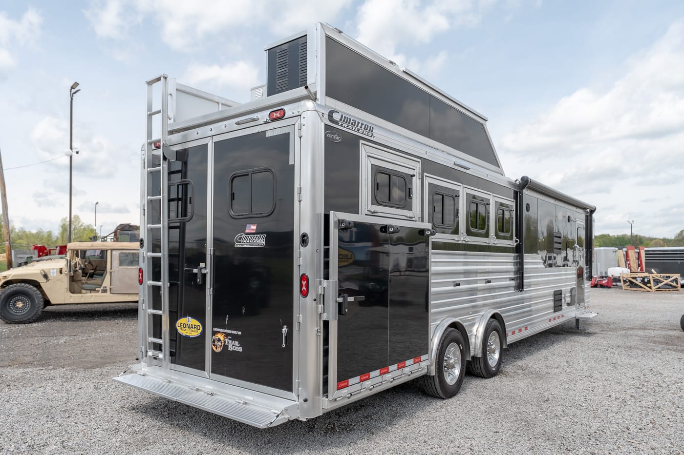 Two-tone black and silver gooseneck horse trailer with living quarters, rear ladder, and multiple side windows parked on a gravel lot.