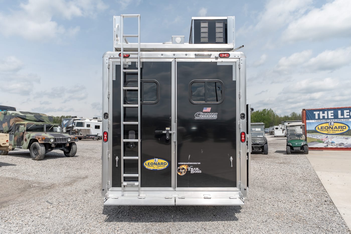 Rear view of a black cargo trailer with a metal ladder on the left, parked on a gravel lot with other vehicles nearby.