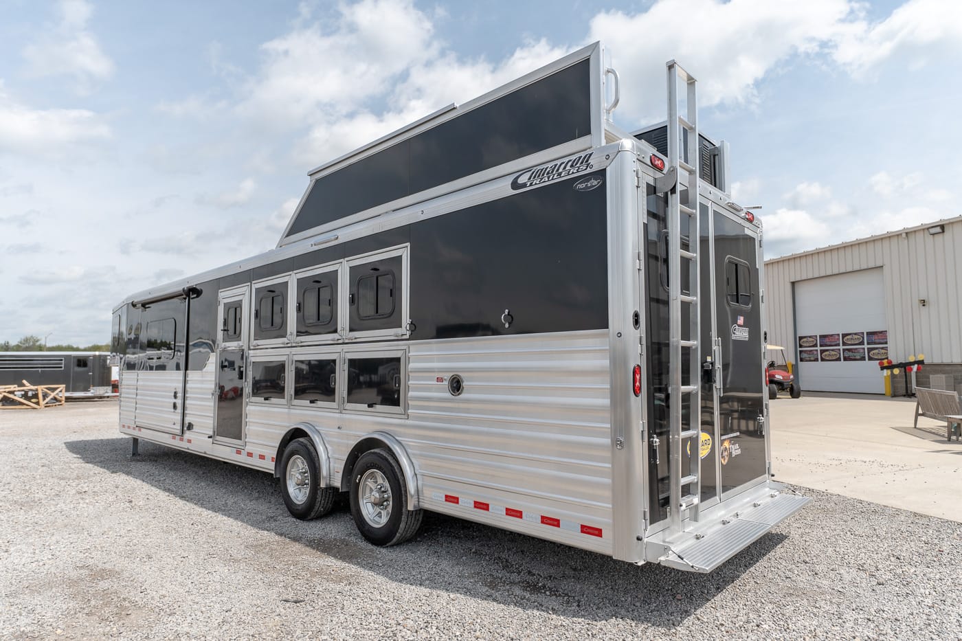 Large two-tone aluminum horse trailer on a gravel lot, with multiple stall windows and a rear ramp.