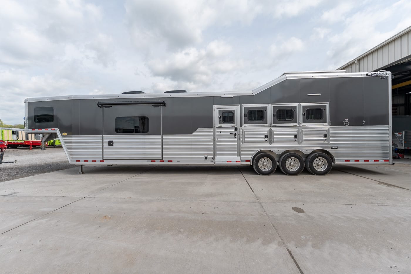 Long gray aluminum horse trailer with multiple stalls and three axles parked on a concrete lot under a cloudy sky