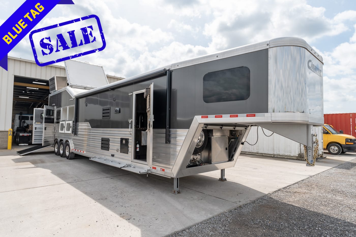 Large silver gooseneck trailer with side access doors parked outside a warehouse, and a blue 'BLUE TAG' SALE banner in the corner.