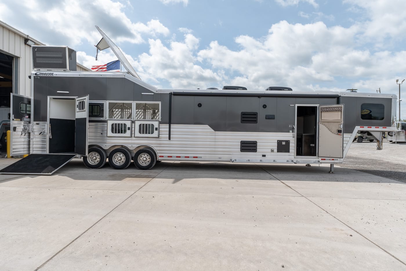 Large gray and silver horse trailer with living-quarters inside, six wheels, open rear ramp, and an American flag in a sunny, cloudy sky.