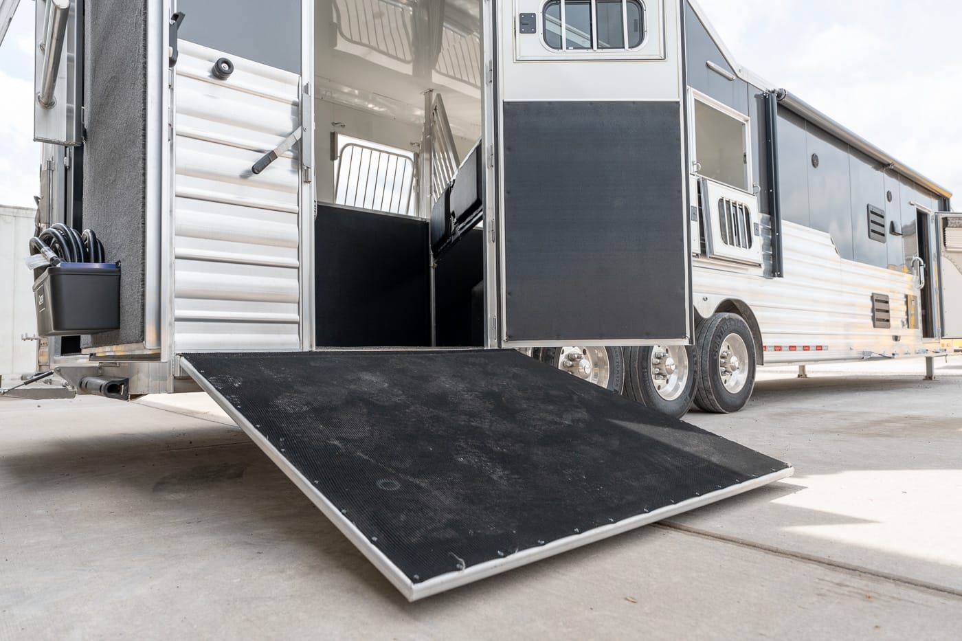 Open horse trailer with the rear ramp down and interior stalls visible on a concrete surface, multiple axles underneath.
