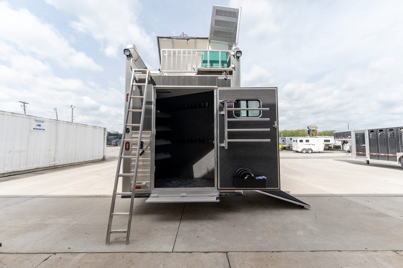 Back of a large metal trailer opened to reveal an interior storage area with shelves; a ladder leans against the side and a ramp is extended on concrete in a truck yard.