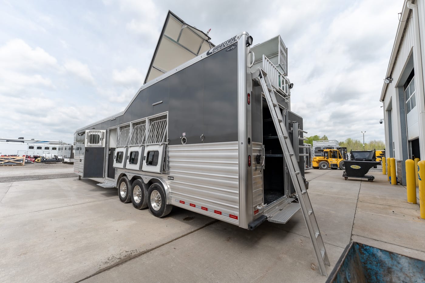 Aluminum horse trailer with multiple stalls and a rear loading ramp parked in a service yard, ladder leaning against it.
