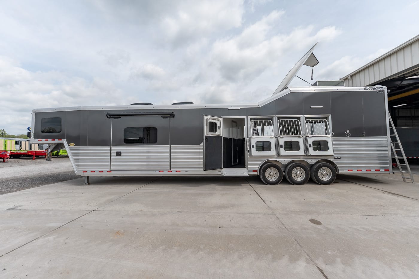 Long silver and gray horse trailer with three axles parked on a concrete lot; stalls visible through open doors on the side.