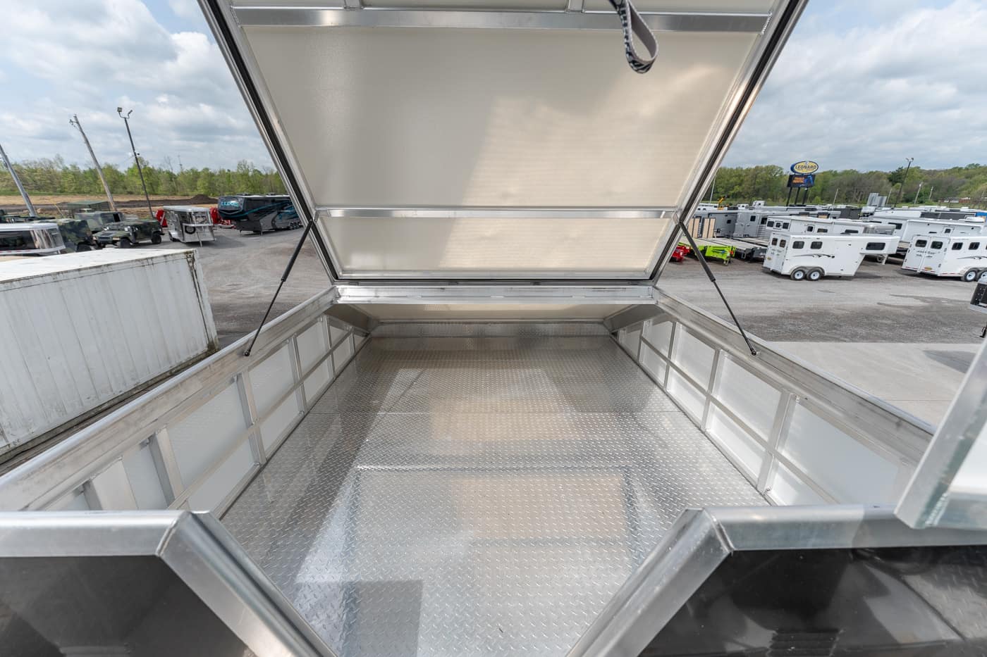 Open aluminum cargo trailer with a diamond-plate floor and raised lid, parked in a lot of horse trailers and RVs at a dealership campground.