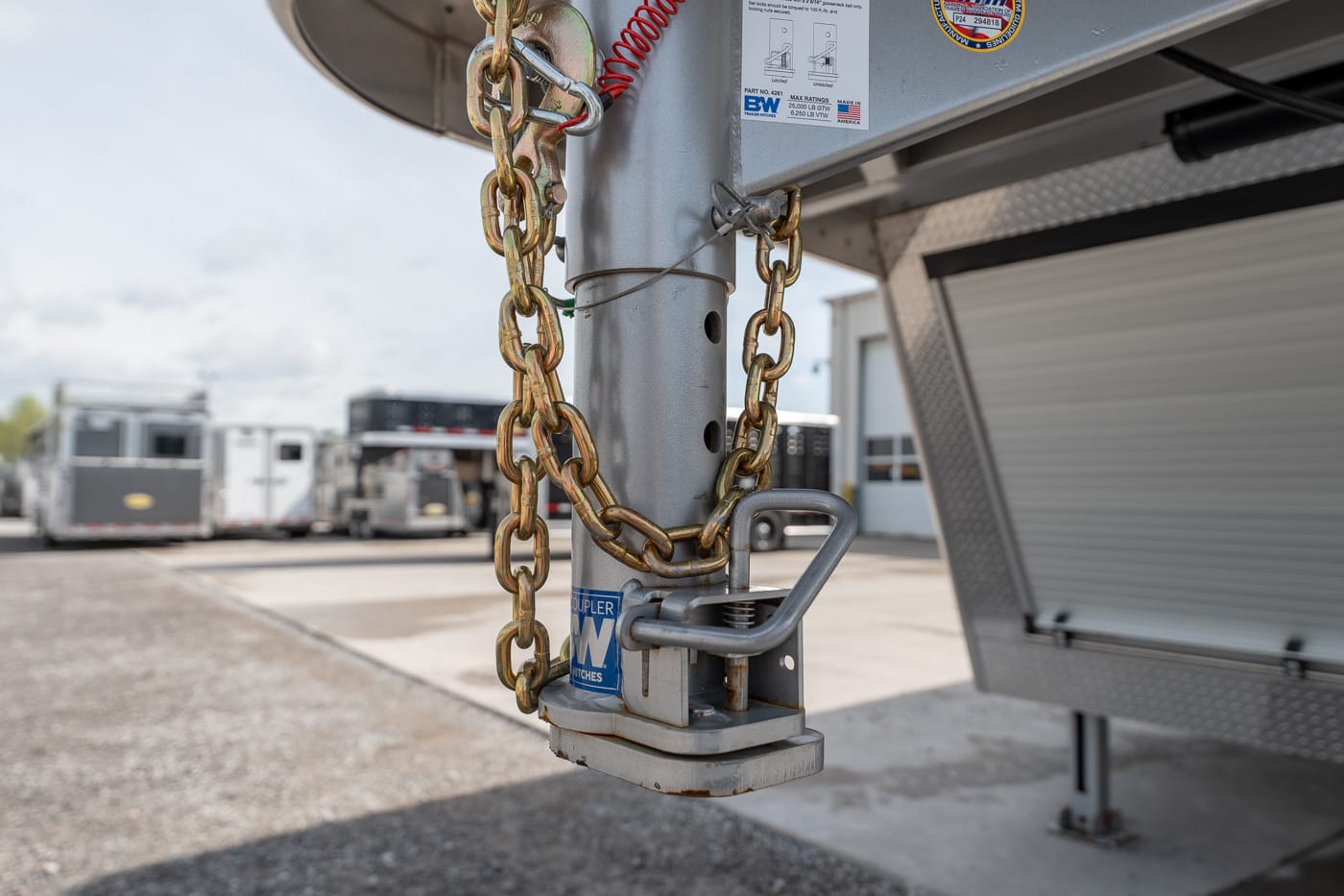 Close-up of a trailer hitch on a silver trailer, secured with a heavy chain and padlock on a gravel lot.
