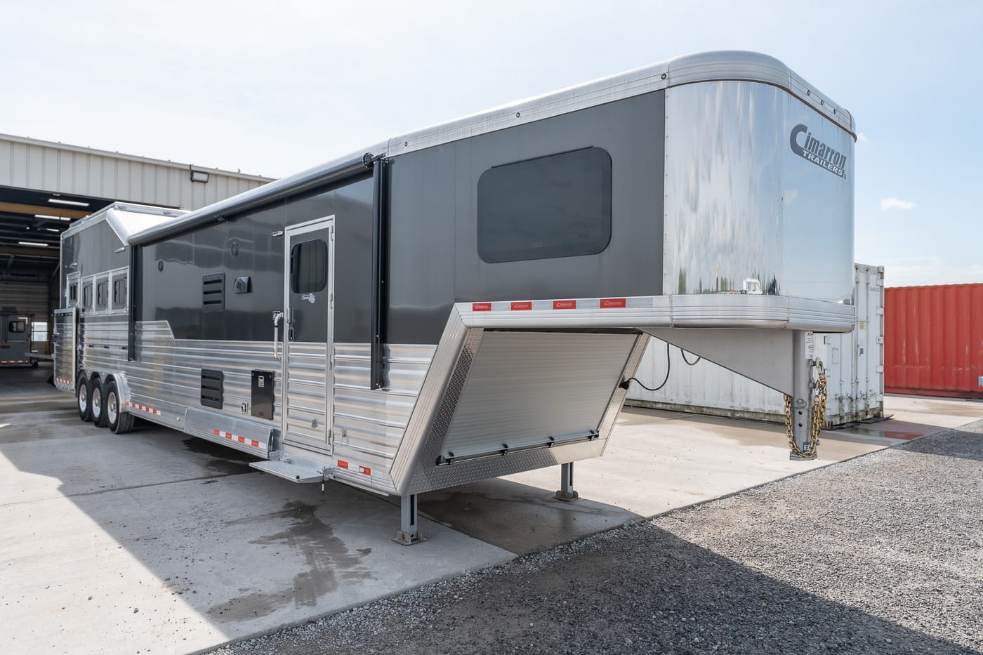 Silver gooseneck horse trailer with raised front compartment and ramp down, parked on a concrete pad near a warehouse site.