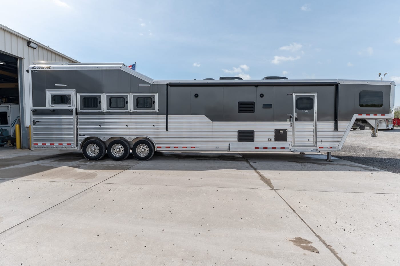 Long aluminum horse trailer with three axles parked outside, gray upper section and silver lower panel.