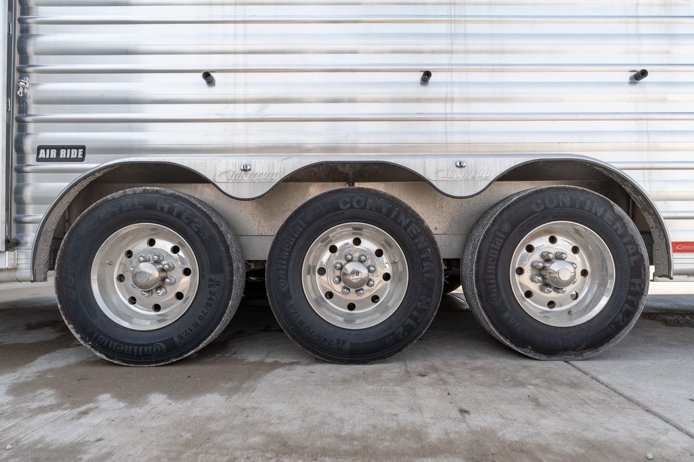 Side view of a metal trailer with three large wheels and hubcaps, showing heavy-duty tires and wheel wells under the body.