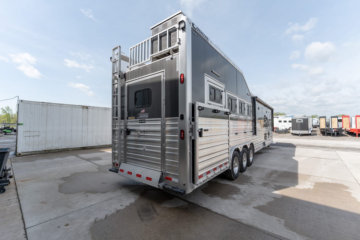Large silver multi-axle horse trailer parked in an outdoor lot, with other trailers visible in the background on a sunny day.