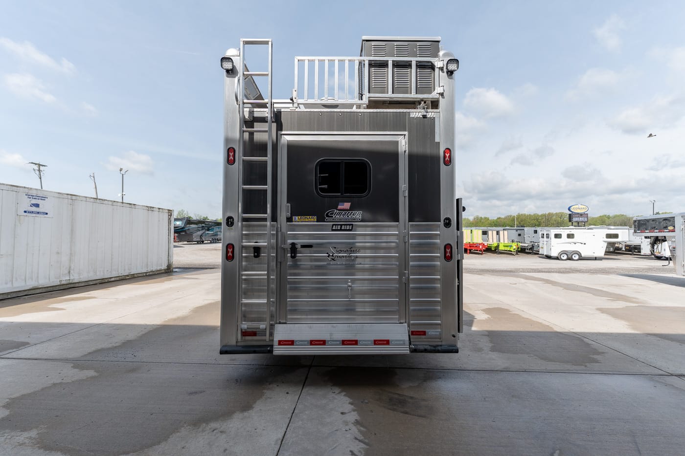 Rear of a shiny aluminum trailer with a small window and ladder on the left, parked in a lot with other trailers.