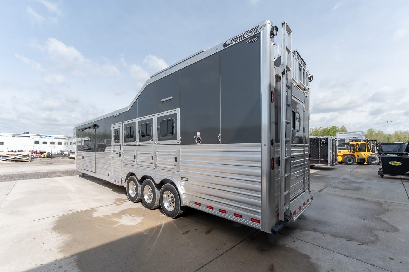 Long aluminum horse trailer with three axles and multiple windows, parked in a truck yard with equipment in the background.
